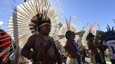 Três homens indígenas adornados com grandes cocares redondos de penas em suas cabeças, segurando seus maracás